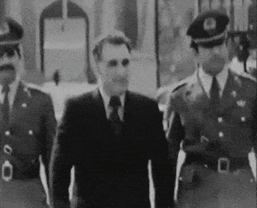 a black and white photo of a man in a suit and tie walking with two other men in uniform .