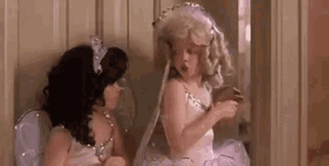 two little girls dressed up as fairy dolls are sitting next to each other in a hallway .
