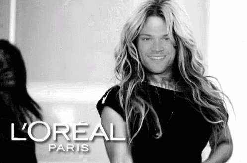 a man with long blonde hair is wearing a black dress and smiling in a black and white photo for loreal paris .