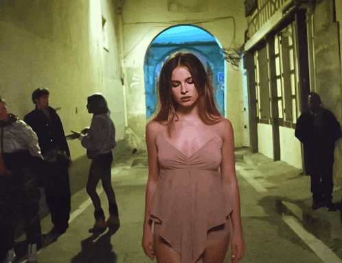 a woman in a pink dress stands in a dark alleyway surrounded by people