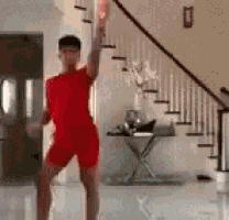 a man in a red shirt and shorts is dancing in a living room with stairs in the background .