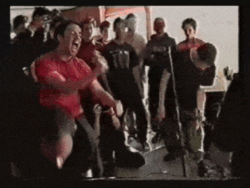 a group of people are standing around a microphone while a man in a red shirt screams into it .