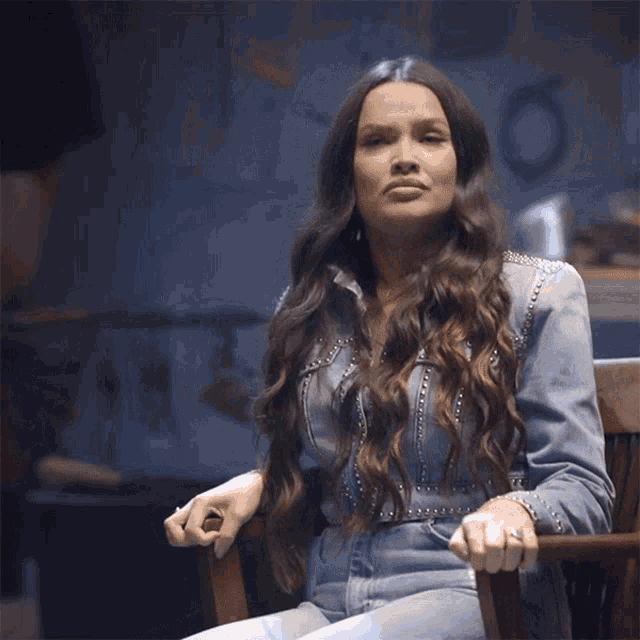 a woman with long hair is sitting in a chair wearing a denim jacket