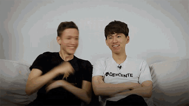 a man wearing a descente shirt sits next to another man on a couch