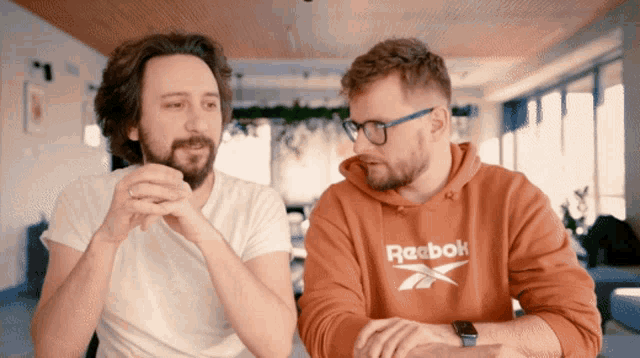 a man wearing a red reebok sweatshirt talks to another man