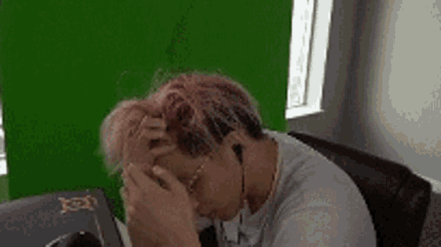 a man with pink hair is sitting at a desk with his hands on his face .