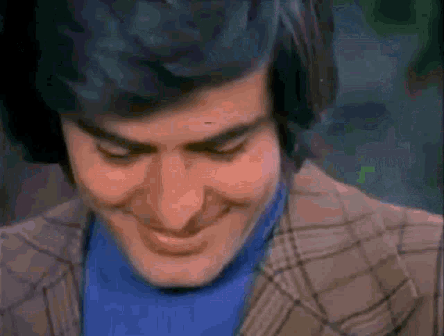a man wearing a plaid jacket and a blue sweater is smiling with his eyes closed .