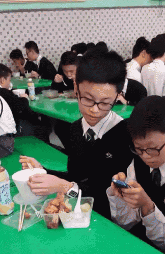 a group of young boys are sitting at green tables eating and looking at their cell phones