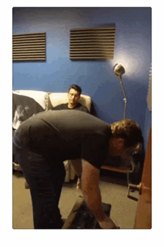a man in a black shirt is standing in front of a door while another man sits on a couch behind him