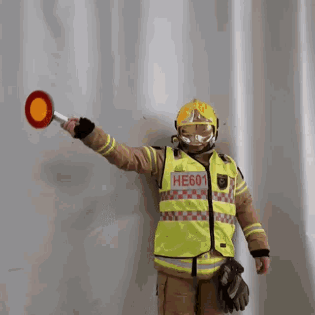 a fireman in a yellow vest with the number he601 on it is holding a red sign .