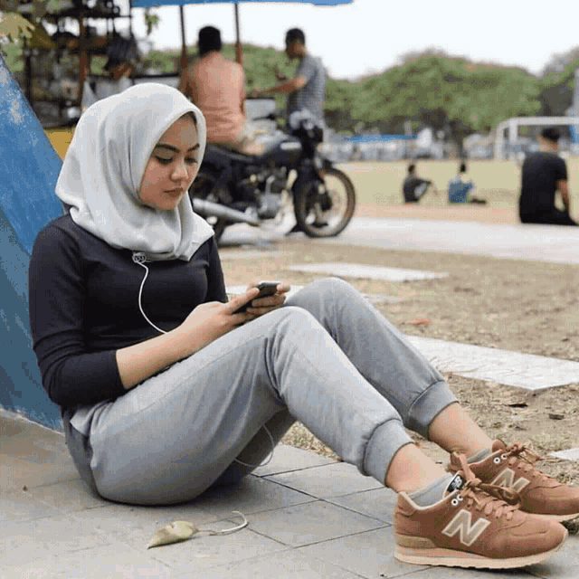 a woman wearing a hijab and new balance shoes sits on the ground looking at her phone