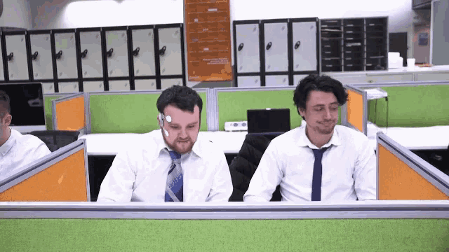 two men are sitting in an office cubicle and one has a sticker on his forehead that says ' a '
