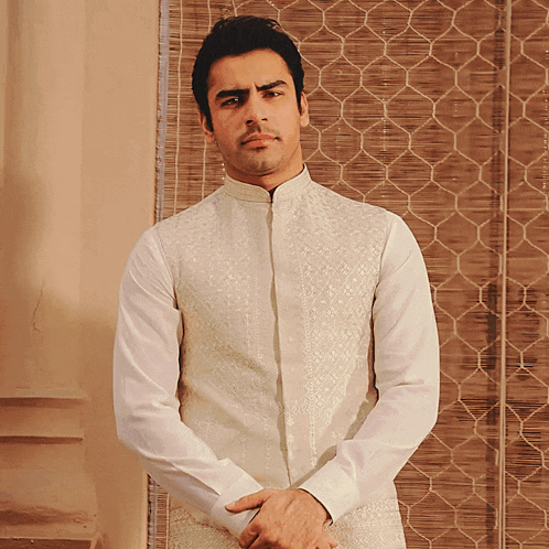 a man in a white shirt stands with his hands folded in front of a blind
