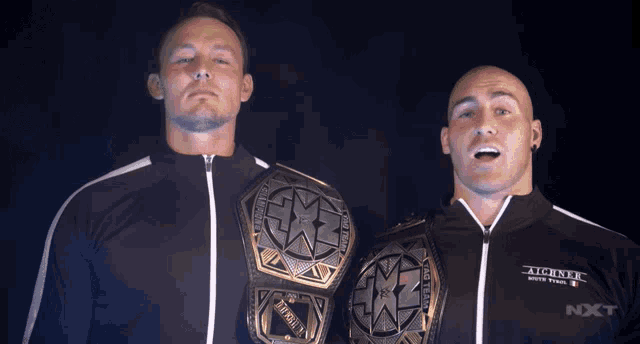 two wrestlers standing next to each other with one wearing a jacket that says nxt on it