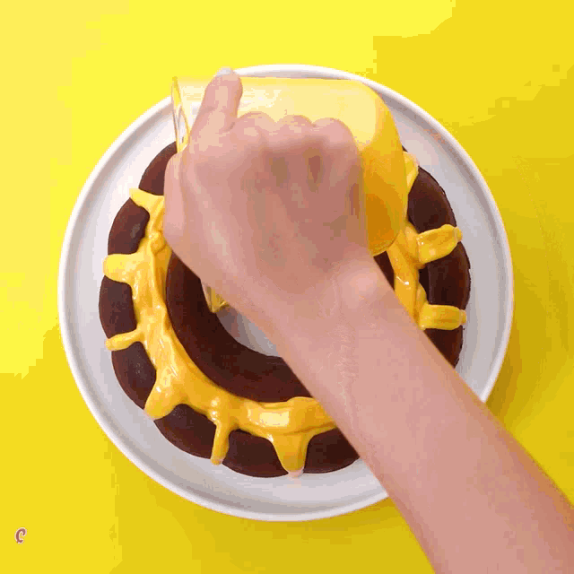 a person pouring yellow frosting on a chocolate cake on a white plate