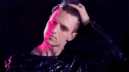 a man in a black shirt is touching his hair in a dark room with pink lights behind him .