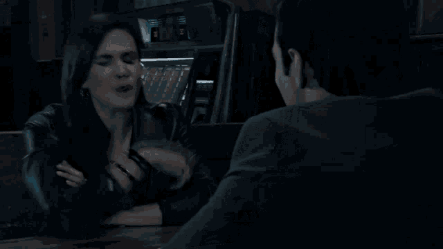 a woman in a leather jacket smiles while talking to a man in a dark room