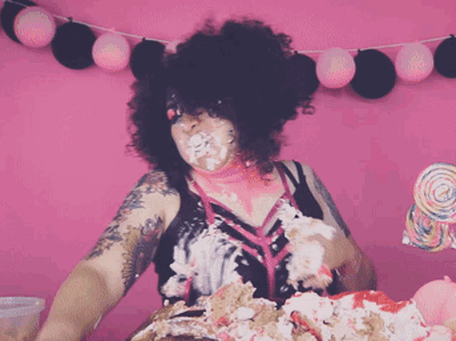 a woman with a lot of cake on her face is standing in front of a pink background