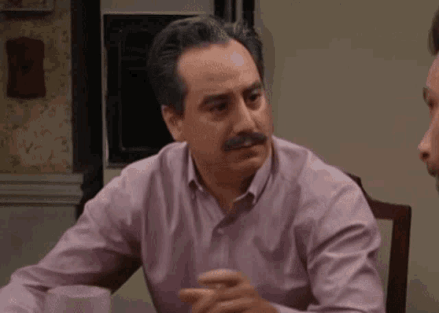 a man in a pink shirt with a mustache is sitting at a table with another man .