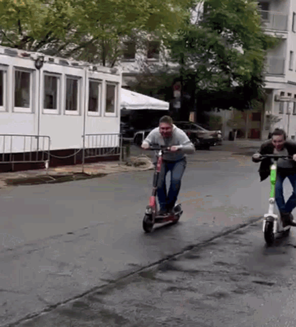 two men are riding scooters down a street in front of a building