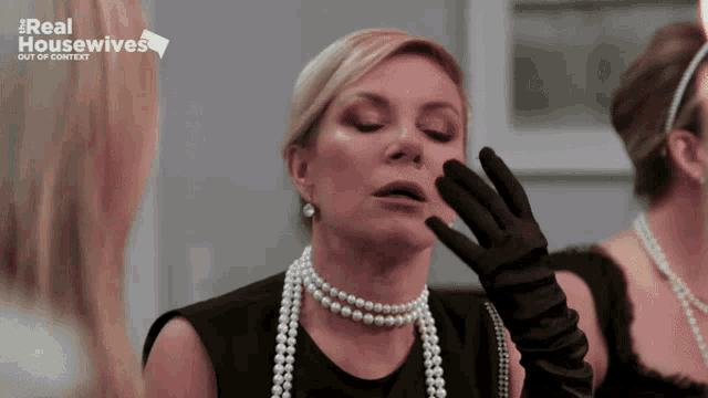 a woman wearing a pearl necklace and black gloves looks at herself in the mirror