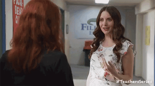 two women are standing next to each other in a hallway talking to each other .