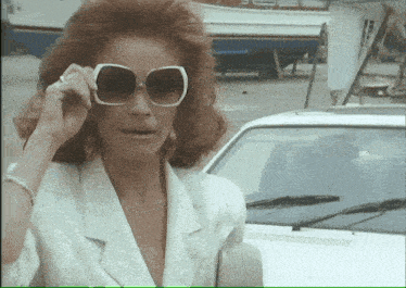 a woman adjusts her sunglasses in front of a car