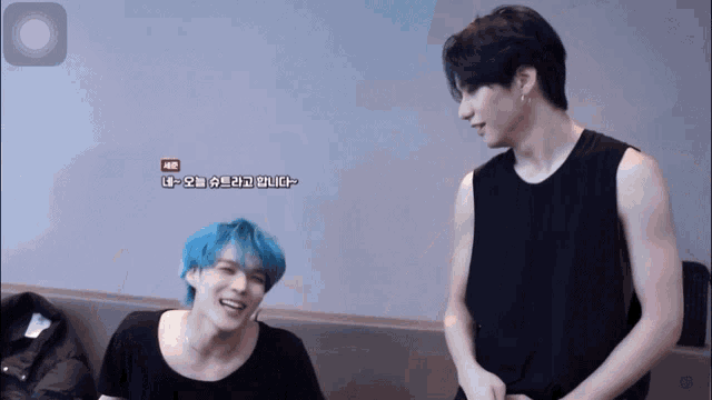a man with blue hair talks to another man in a black tank top