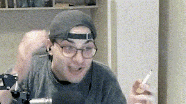 a man wearing a hat and glasses is holding a cigarette