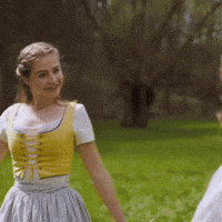 a woman in a yellow top and gray skirt is standing in the grass holding hands with a man .
