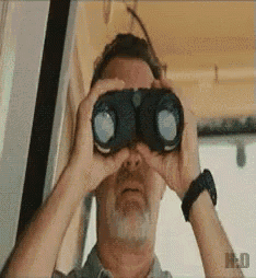 a man with a beard is looking through binoculars with the letter h on the bottom right
