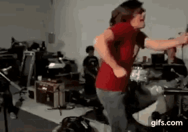 a man in a red shirt is dancing in a room with a drum set in the background .