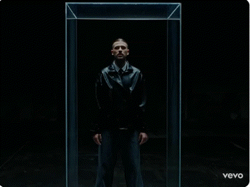 a man in a black jacket is standing in a glass box with his hands on his chest .