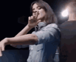 a woman in a denim shirt is giving a thumbs up in a dark room .
