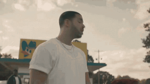 a man is standing in front of a gas station wearing a white shirt and a necklace .
