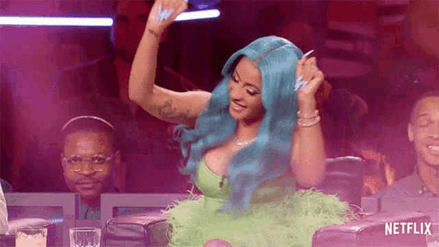 a woman with blue hair is sitting at a table with her arms in the air and smiling .