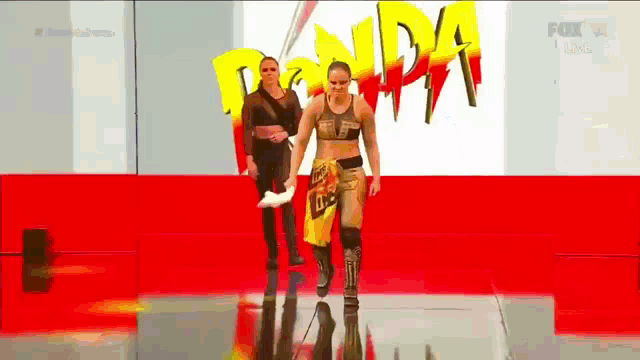 two women are walking on a red carpet in front of a sign that says ' ronda rousey ' on it .