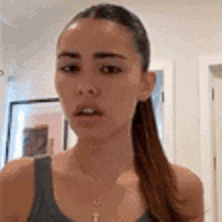 a woman in a tank top and ponytail is standing in a room and looking at the camera .