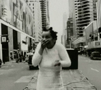 a black and white photo of a woman standing on a city street covering her mouth with her hand .