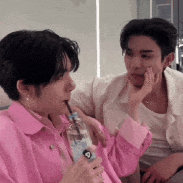 a man in a pink jacket is drinking water through a straw while another man in a white shirt looks on .