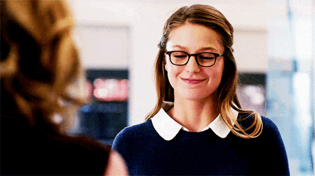 a woman wearing glasses and a blue sweater smiles while talking to another woman