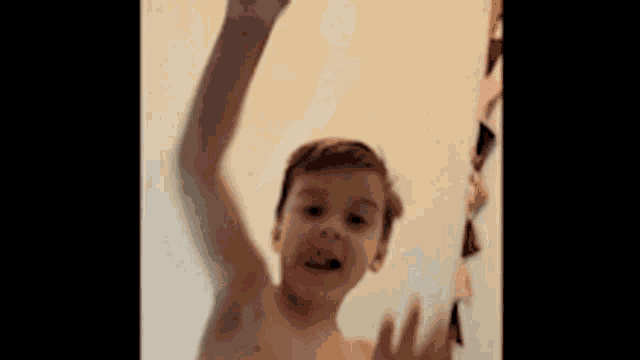 a young boy without a shirt is standing in front of a wall with his hand up .