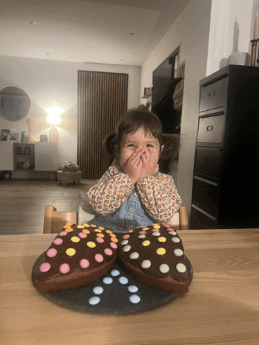 a little girl covering her mouth with her hands while sitting at a table with two chocolate hearts on it