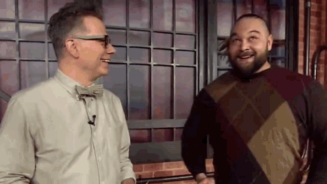 two men are standing next to each other in front of a window and smiling . one of the men is wearing a bow tie .