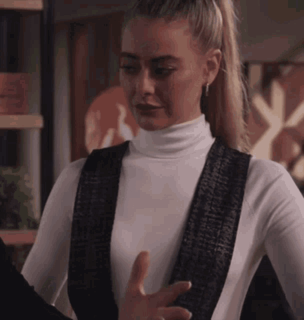 a woman wearing a white turtleneck and black overalls has her hand out