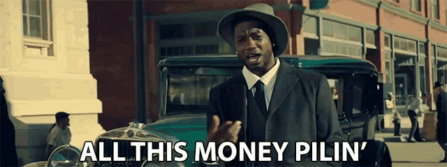 a man in a suit and hat is standing in front of a car and says all this money pilin '