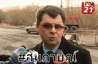 a man wearing glasses and a suit is talking into a microphone while a car drives in the background .