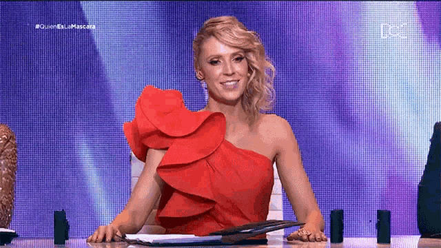 a woman in a red dress is sitting at a table with a clipboard and smiling .