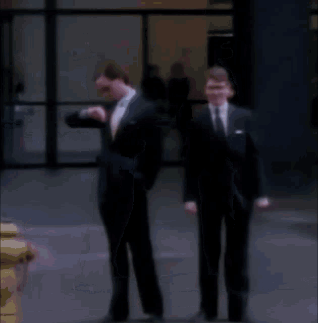 two men in suits shake hands on a sidewalk in front of a building