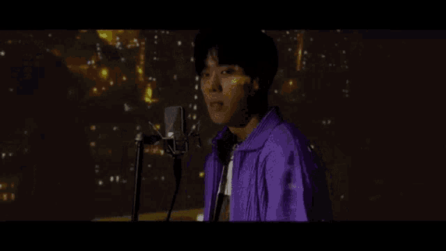 a man in a purple jacket is singing into a microphone in front of a city at night .